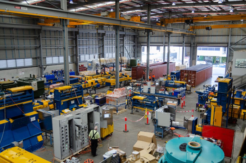 An industrial warehouse with large machinery and equipment. Workers in safety vests move around the space, which is filled with colorful machines, shipping containers, and various tools. Overhead cranes are visible above the workspace.