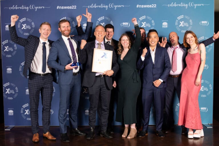 A group of ten people in formal attire celebrate together in front of a blue backdrop with "celebrating 60 years" and "#nswea22" logos. One person holds an award, and they all have joyful expressions, some raising their fists in excitement.