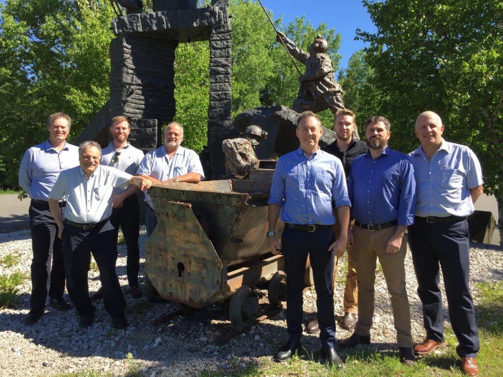 A group of men stands outdoors in front of a rusted mining cart and stone monument, surrounded by green trees. They are all dressed in business casual attire, smiling under a clear blue sky.