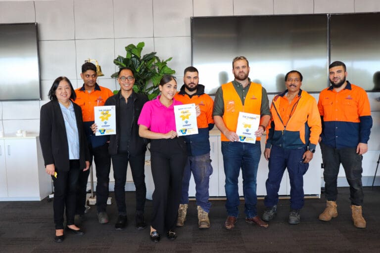 A group of eight people stands indoors, smiling. Some hold certificates. Four individuals wear orange work uniforms, while others are in casual or formal attire. Two large screens and a potted plant are visible in the background.