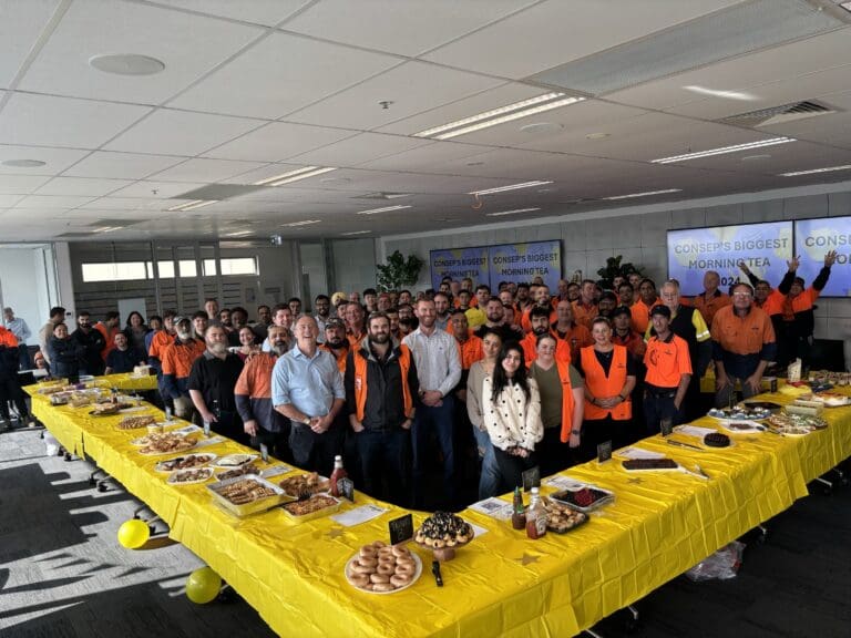 A large group of people gather in an office setting, standing around tables covered with yellow cloths and a variety of foods. There are balloons and signs that read "CONSEP'S BIGGEST MORNING TEA 2023" in the background.