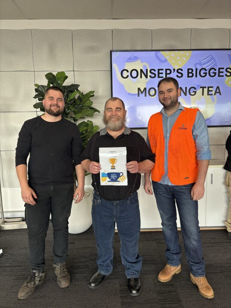 Three men stand together, smiling in front of a screen displaying the words "Consep's Biggest Morning Tea." The man in the center holds a certificate with a trophy image. One man wears an orange safety vest. A potted plant is visible in the background.