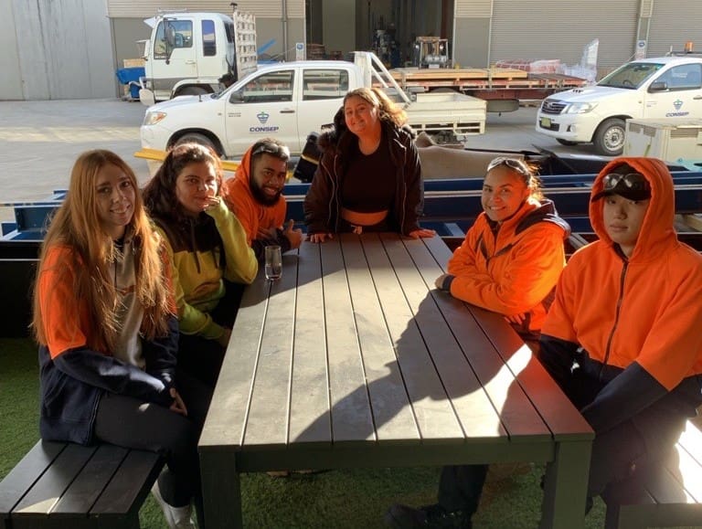 A group of six people wearing casual and high-visibility clothing sit around a rectangular table outdoors. They are in a sunny area near industrial buildings and parked vehicles, enjoying a break.