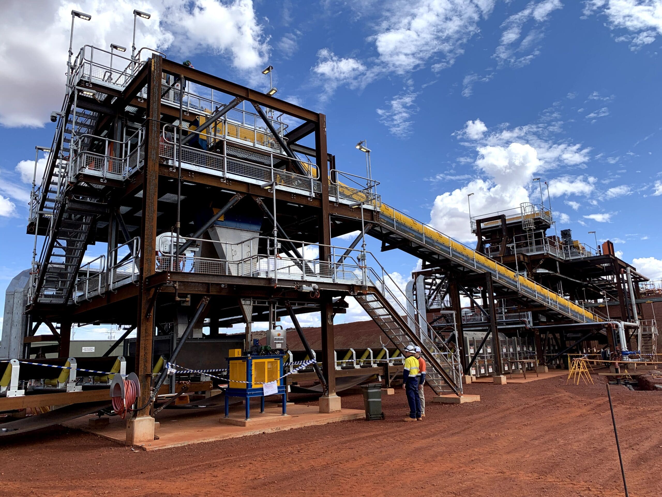 A large industrial structure with metal frameworks and conveyor belts set against a blue sky with clouds. Two workers in safety gear stand at the base on reddish-brown ground, emphasizing the site's construction and manufacturing elements.