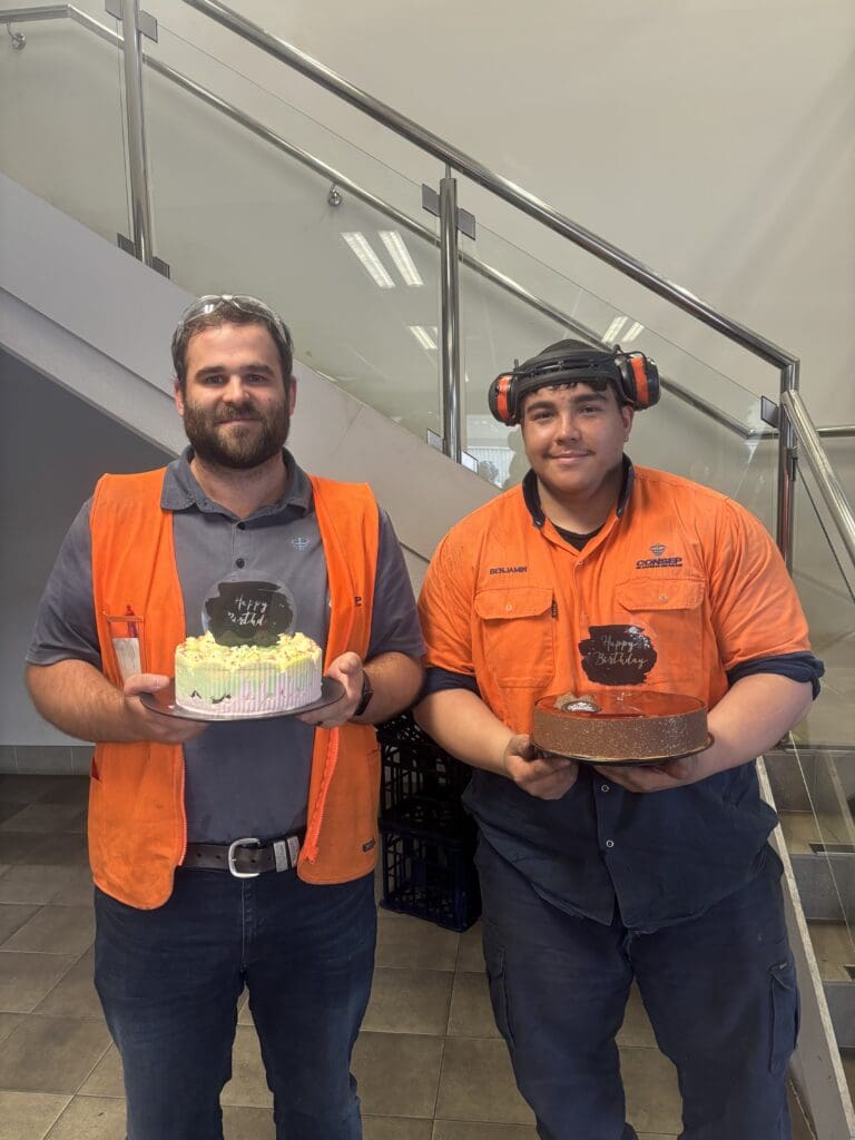 Two people wearing orange safety vests are standing indoors. They are smiling and each holding a cake. They are positioned in front of a staircase.