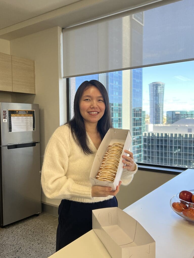 A person in a white sweater smiles while holding an open pastry box with a pastry inside. They stand in a kitchen area with a window showing tall buildings in the background. A bowl of apples is on the counter.