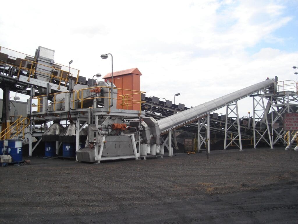 Image of an industrial site featuring a conveyor system with multiple belts and metal structures. The setup is supported by steel framework, and there's a large cylindrical tube leading upwards. The ground is covered in gravel. Sky is overcast.
