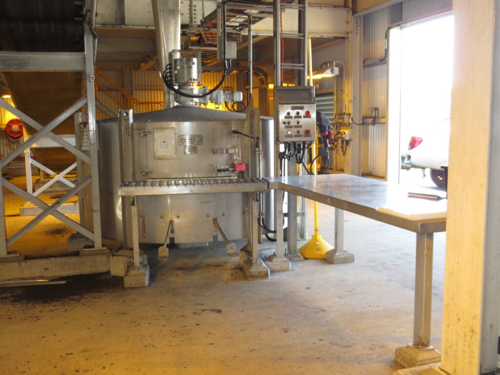 Industrial setting with a large metal machine connected to pipes. The machinery has control panels and a conveyor belt leading to an opening on the right. The room is lit by natural light from a door opening.