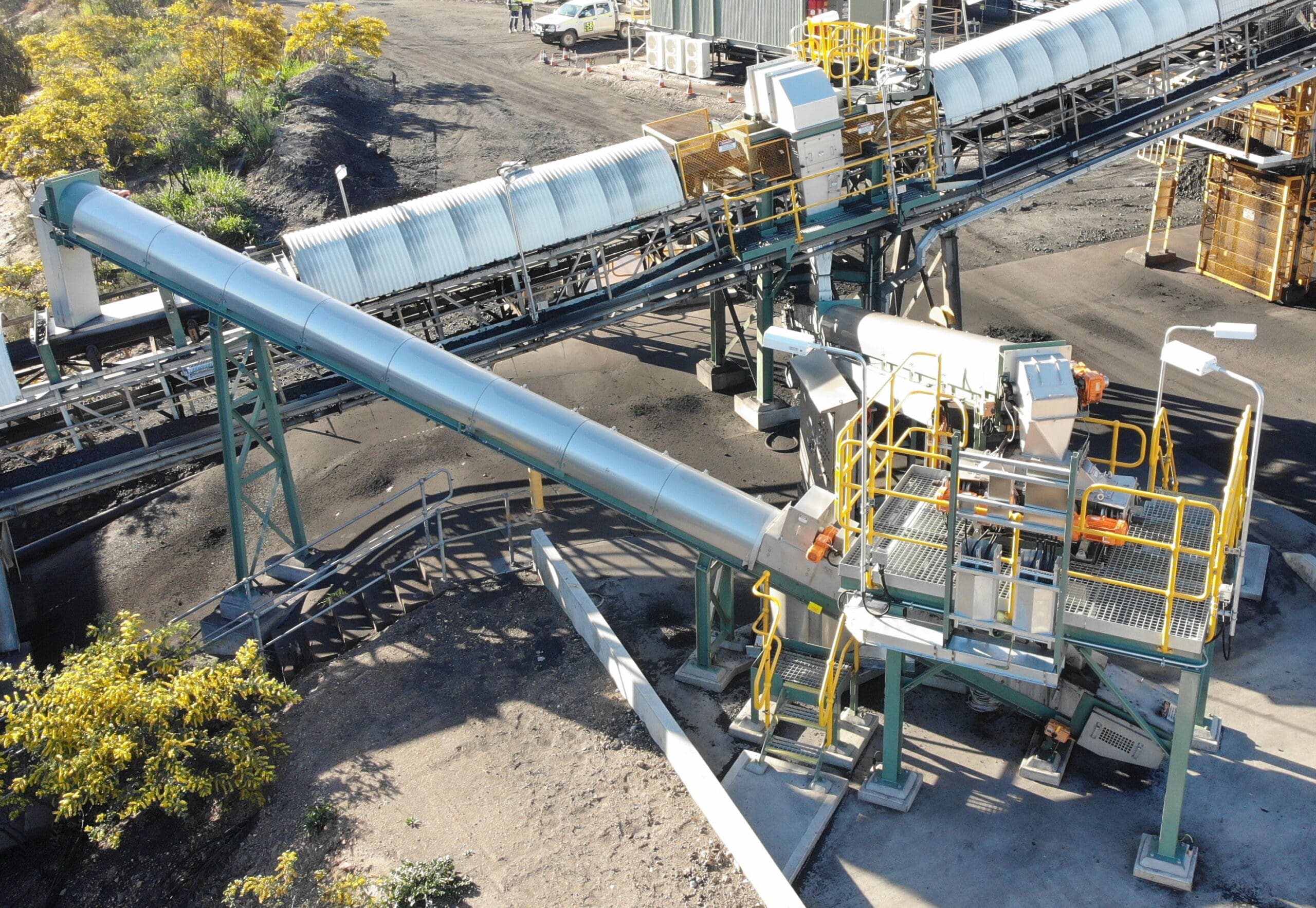 Aerial view of an industrial site with conveyor belts transporting materials. The structure includes metal beams, safety railings, and staircases, surrounded by a mix of greenery and industrial equipment on the ground.