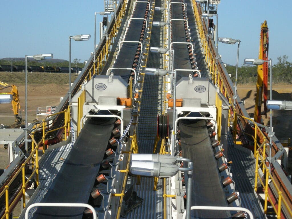 A conveyor belt system at an industrial site, with multiple parallel belts carrying materials. The machinery features metal supports and orange rollers, surrounded by industrial equipment and a clear blue sky in the background.