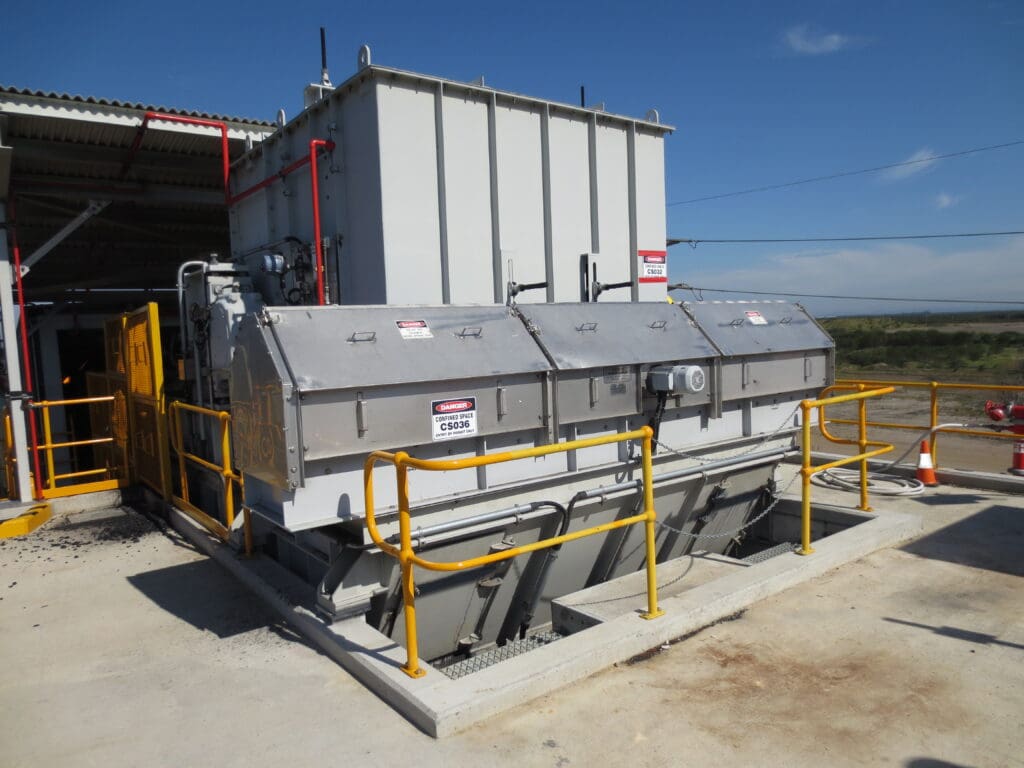 Large industrial equipment outdoors with metal housing, safety railings, and signs. The machinery is on a concrete platform with railings and cones nearby. There is a clear sky and open landscape in the background.